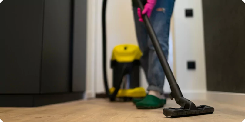 Staff member vacuuming wooden flooring in a corridor of a remote camp facility with yellow cleaning equipment.