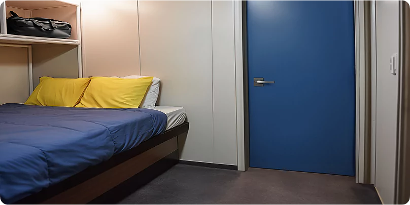 Single room in a workforce camp with a bed, blue bedding, yellow pillows, built-in shelving, and a blue door.