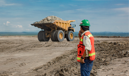 Worker radios colleague at mine site with dump truck in background