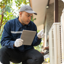 Maintenance worker holds tablet and studies air conditioner outside in tropical setting.