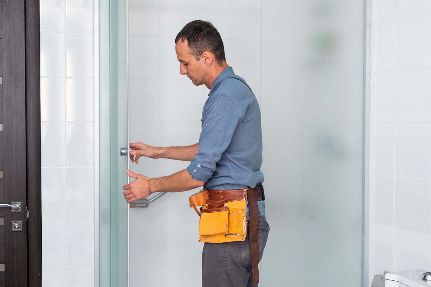 Maintenance staff fixes a shower door, wearing a toolbelt.